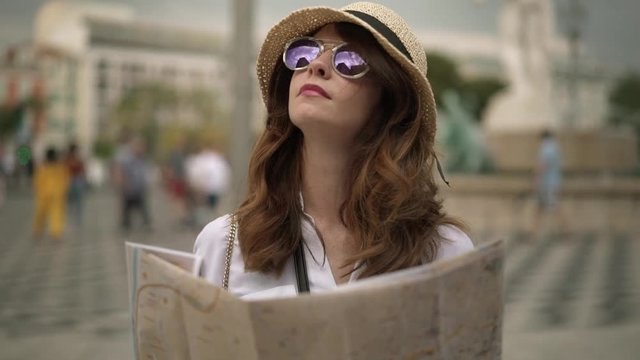 Beautiful caucasian woman tourist with long brown hair wearing a hat and purple sunglasses is standing in blurred street of Nice, France with a map and looking around. Slider slow motion medium shot