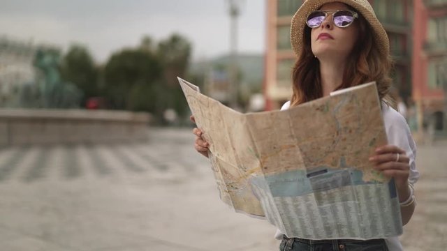 Young woman tourist with long brown hair wearing a hat and sunglasses is standing in street of Nice, France on cloudy day holding a map and looking around. Left to right pan slow motion medium shot
