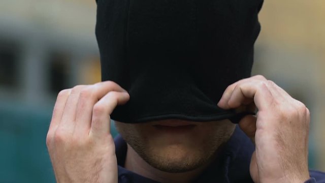 Aggressive criminal puts on balaclava and hood hiding face before attack, danger