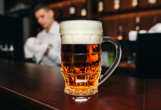 Glass of beer with foam is standing on the wooden bar counter. Old pub with bartender cleaning the dishes on background. Leisure, fun and friends concepts photo.