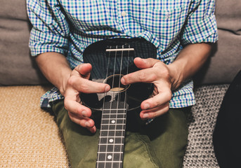 Man in checkered shirt at home is playing the ukulele guitar. Guys hands are taking the chord on...