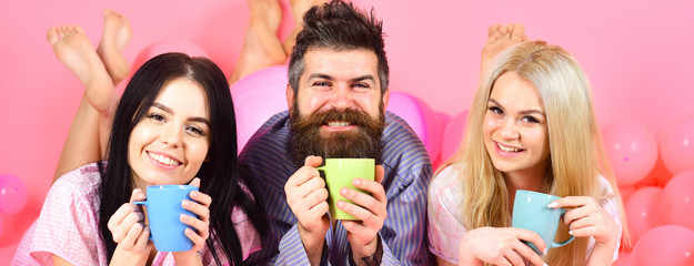 Man and women in domestic clothes, pajamas. Threesome relax in morning with coffee. Stormy night concept. Man and women, friends on smiling faces lay, pink background. Lovers drinking coffee in bed.