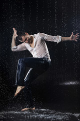 Fototapeta na wymiar A young barefoot man wearing wet blue jeans and a white shirt expressively dances modern dances on the water under rainy waterdrops among the watersplashes