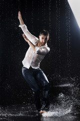 Fototapeta na wymiar A young barefoot man wearing wet blue jeans and a white shirt expressively dances modern dances on the water under rainy waterdrops among the watersplashes