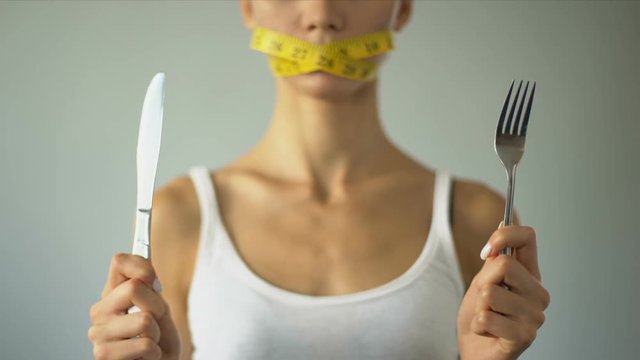 Slim girl closed mouth with tape, holding fork and knife, excessive self-control