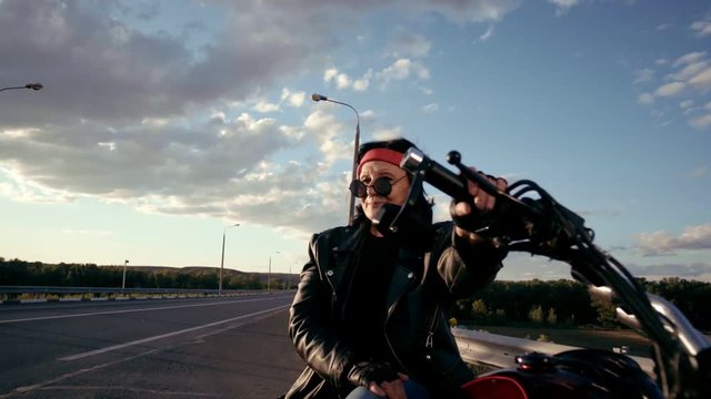 Old woman biker in a leather jacket and gloves sitting on his motorcycle. There's an empty highway in the background. The life of an elderly rocker and biker after retirement. A woman in round glasses