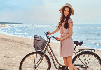 Beautiful brunette girl dressed in dress and hat posing with a bicycle on the beach on a sunny day.