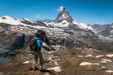 Fototapeta premium Female traveler with backpack hiking mountain trail and admiring views of Majestic Matterhorn mountain in Valais (Pennine) Alps, Switzerland.