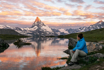 Obraz premium Morning shot of the golden Matterhorn (Monte Cervino, Mont Cervin) pyramid and blue Stellisee lake. Female tourist enjoying view of early morning Matterhorn mountain is Valais Alps, Switzerland.