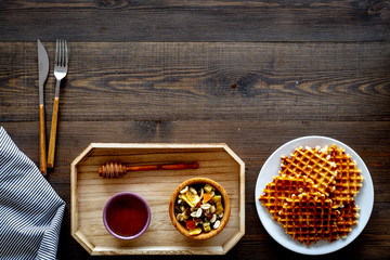 Round belgian waffles for breakfast. Breakfast in bed. Waffles on plate. Honey and dried fruits in tray, knife and fork, tablecloth on dark wooden background top view copy space