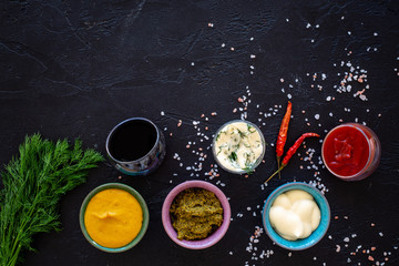 Different dip sauce in bowls near chili pepper and greenery. Ketchup, mayonnaise, mustard, soy sauce, barbecue sauce, pesto, mustard, sour on black background top view copy space