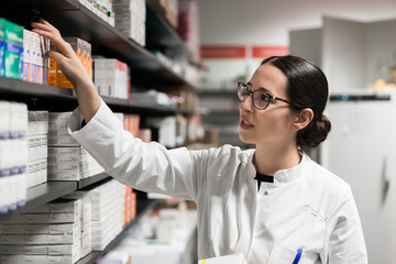 Portrait of a dedicated female pharmacist taking a medicine from the shelf, while wearing eyeglasses and lab coat during work in a modern drugstore with various pharmaceutical products