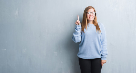 Young adult woman over grey grunge wall wearing glasses showing and pointing up with finger number one while smiling confident and happy.