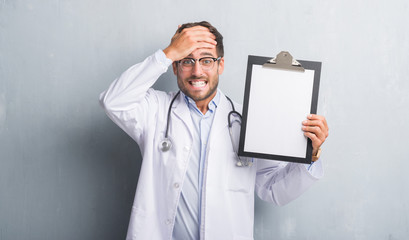 Handsome young doctor man over grey grunge wall holding clipboard stressed with hand on head, shocked with shame and surprise face, angry and frustrated. Fear and upset for mistake.