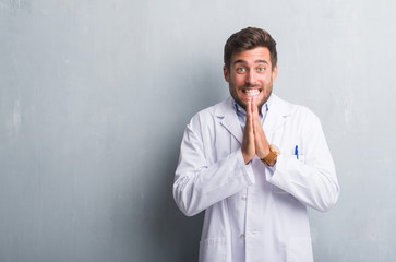 Handsome young professional man over grey grunge wall wearing white coat praying with hands together asking for forgiveness smiling confident.