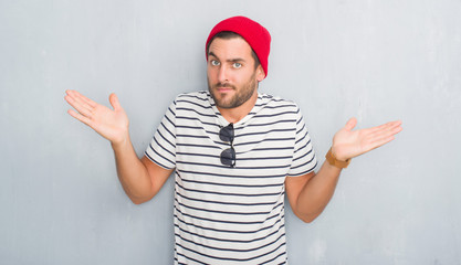 Naklejka premium Handsome young hipster man over grey grunge wall wearing navy t-shirt and wool cap clueless and confused expression with arms and hands raised. Doubt concept.