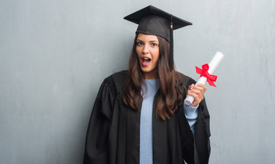 Young brunette woman over grunge grey wall wearing graduate uniform holding degree scared in shock...