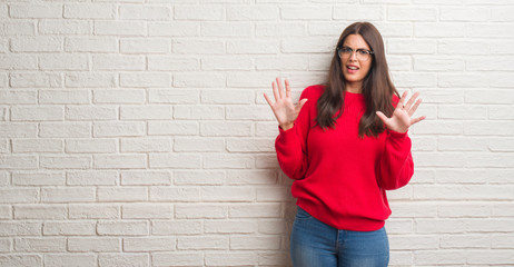 Obraz na płótnie Canvas Young brunette woman standing over white brick wall afraid and terrified with fear expression stop gesture with hands, shouting in shock. Panic concept.