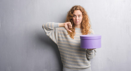 Young redhead woman over grey grunge wall holding box with angry face, negative sign showing dislike with thumbs down, rejection concept