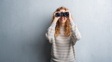 Young redhead woman over grey grunge wall looking through binoculars with a happy face standing and...