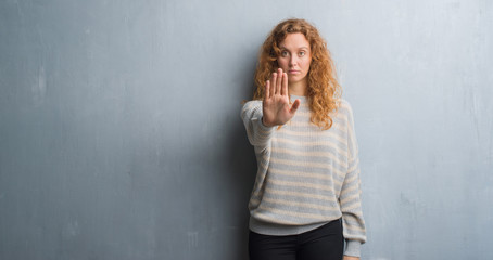Young redhead woman over grey grunge wall doing stop sing with palm of the hand. Warning expression with negative and serious gesture on the face.