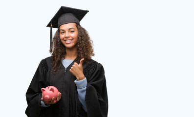 Young hispanic woman wearing graduation uniform holding piggy bank happy with big smile doing ok sign, thumb up with fingers, excellent sign