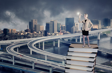 A businesswoman standing on a stack of books looking through spyglass against a city downtown