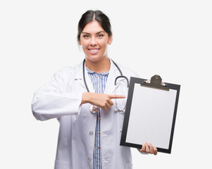 Young hispanic doctor woman holding clipboard very happy pointing with hand and finger