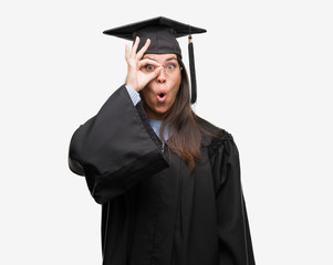 Young hispanic woman wearing graduated cap and uniform doing ok gesture shocked with surprised face, eye looking through fingers. Unbelieving expression.