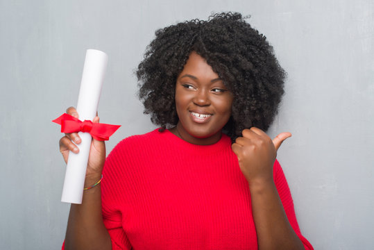 Young african american woman over grey grunge wall holding diploma pointing and showing with thumb up to the side with happy face smiling