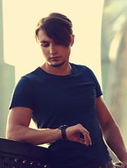 Thinking handsome young man looking on watch on the hand on city center background. Closeup toned...