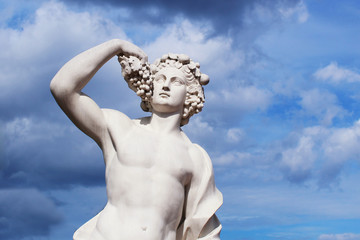 Sculpture of a young guy of white material with grapes against a blue sky and clouds