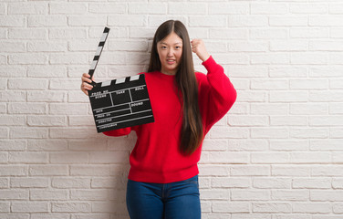 Young Chinese woman over white brick wall holding clapboard annoyed and frustrated shouting with...