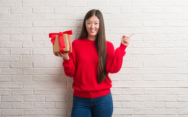 Young Chinese woman over brick wall holding a gift very happy pointing with hand and finger to the side