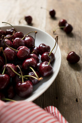 Cherries in a white bowl on a wood table with a white and red napkin