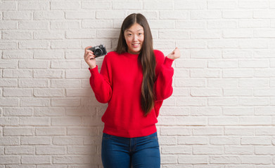 Fototapeta na wymiar Young Chinese woman over brick wall holding vintage camera screaming proud and celebrating victory and success very excited, cheering emotion