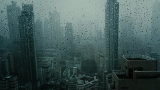 Window with falling rain at dusk in scryscraper.Dark and stormy rainy day in big city life. View of dreary Hong Kong cityscape whilst rain drops fall on the window. Abstract somber moody background.