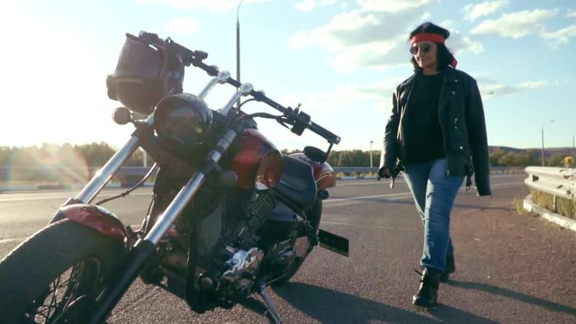 The old biker and his motorcycle. A woman in a leather jacket, a red bandana and wearing round glasses goes to her steep motorcycle. Motorcycle and old biker on a background of nature on an empty
