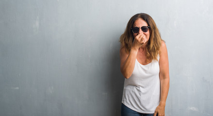 Middle age hispanic woman over grey wall wearing sunglasses smelling something stinky and disgusting, intolerable smell, holding breath with fingers on nose. Bad smells concept.