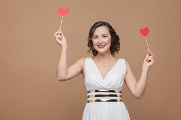 Toothy smiled middle aged elegant brunette woman in white dress standing holding two red heart stickers and looking at camera. Studio shot, indoor, isolated on light brown background