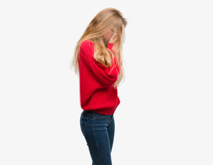 Blonde teenager woman wearing red sweater with sad expression covering face with hands while crying. Depression concept.