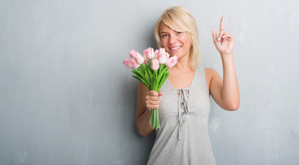 Caucasian adult woman over grey grunge wall holding pink flowers surprised with an idea or question pointing finger with happy face, number one