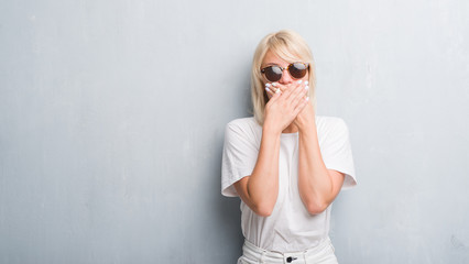 Adult caucasian woman over grunge grey wall wearing sunglasses shocked covering mouth with hands for mistake. Secret concept.