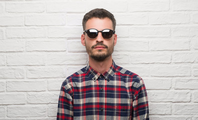 Young adult man wearing sunglasses standing over white brick wall with a confident expression on smart face thinking serious