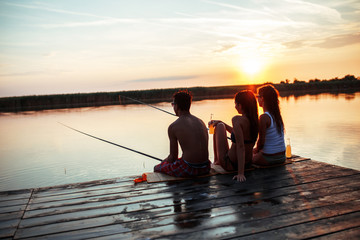 Group of friends sitting on pier by the lake and fishing.They joying in beautiful summer sunset.