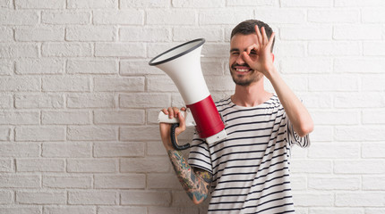 Young adult man holding megaphone with happy face smiling doing ok sign with hand on eye looking through fingers