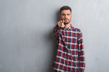 Young adult man standing over grey grunge wall pointing with finger to the camera and to you, hand sign, positive and confident gesture from the front