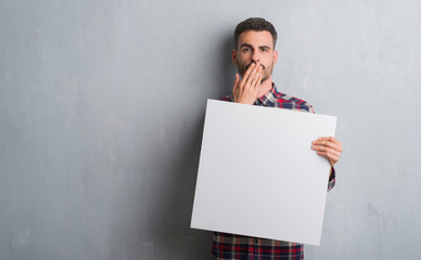 Young adult man over brick wall holding banner cover mouth with hand shocked with shame for mistake, expression of fear, scared in silence, secret concept