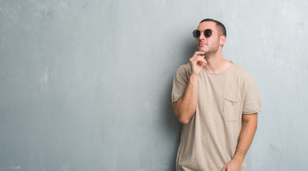 Young caucasian man over grey grunge wall wearing sunglasses with hand on chin thinking about question, pensive expression. Smiling with thoughtful face. Doubt concept.