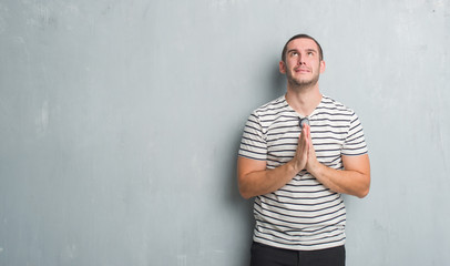 Young caucasian man over grey grunge wall begging and praying with hands together with hope...
